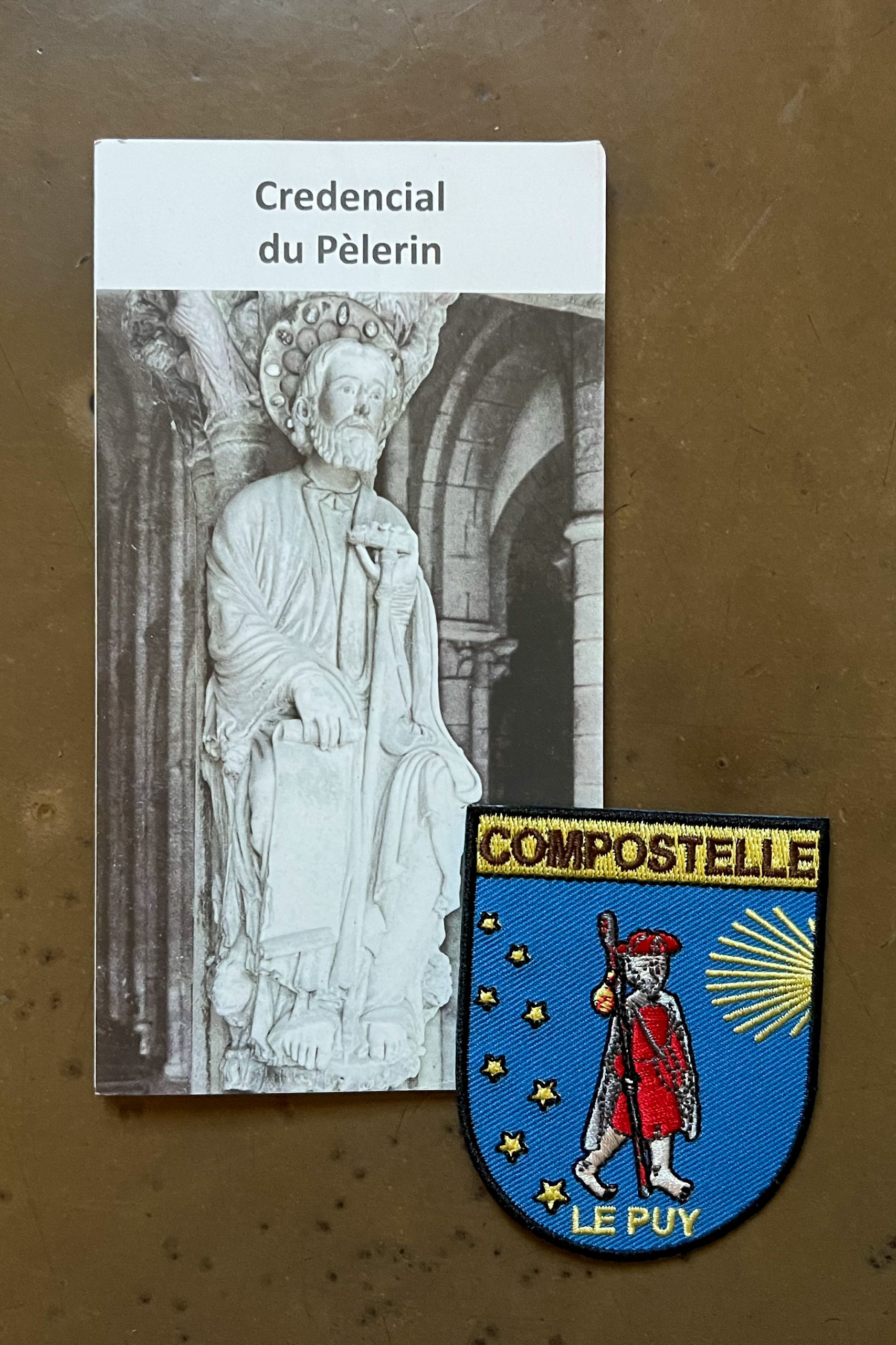 My pilgrim credential and a colony badge that says Compostelle Le Puy and shows a barefoot pilgrim in a red robe and hat and a walking stick