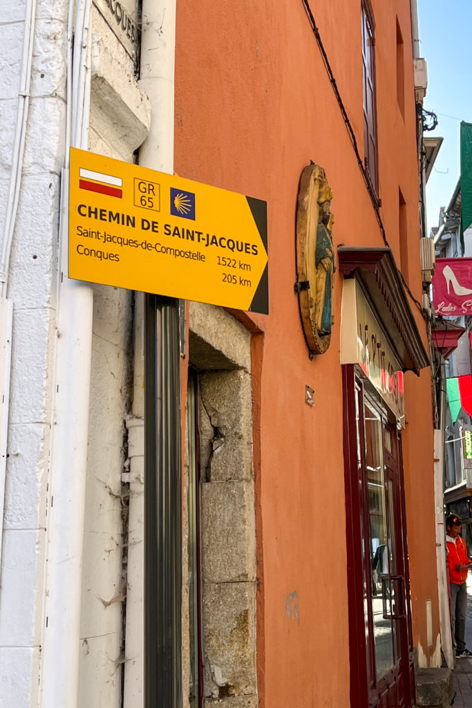 A signpost pointing down a colorful street. The sign says, Chemin de Saint-Jacques, Saint-Jacques-de-Compostelle 1522 km, Conques 205km.
