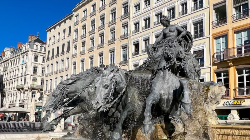 Bartholdi Fountain against a backdrop of old French apartment buildings. The fountain depicts a nude woman in a chariot. She holds the reins of four horses who are galloping before her.